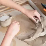 Dłonie formują glinę, tworząc figurki z gliny krok po kroku. Narzędzia ceramiczne i wałek leżą obok.