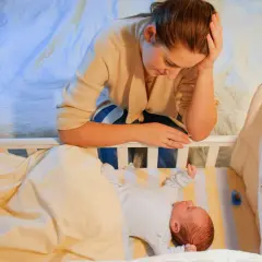 Zmęczona mama pochyla się nad łóżeczkiem, przeżywając kryzys snu u 9-miesięcznego dziecka.