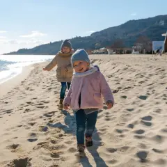Dwa uśmiechnięte dzieci biegają po piaszczystej plaży. W tle widać morze i góry.