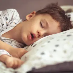 Małe dziecko śpi z otwartą buzią, wtulone w poduszkę. Jego sen jest głęboki i spokojny.