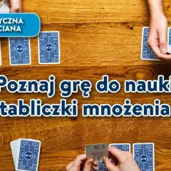 Gra karciana "Poznaj grę do nauki tabliczki mnożenia" na drewnianym stole. Dłonie trzymają karty z działaniami.