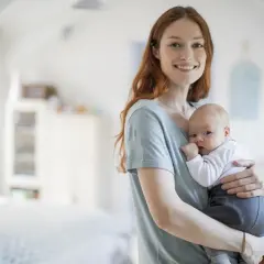 Uśmiechnięta mama trzyma na rękach noworodka. Dziecko ma lekko otwarte usta, jakby właśnie dostało czkawki.