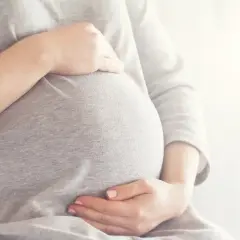 Brzuszek w 6 miesiącu ciąży, otulony szarym materiałem. Dłonie delikatnie go obejmują, czuć radość oczekiwania.
