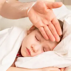 Dłoń rodzica sprawdza czoło śpiącego dziecka. Warto wiedzieć, od jakiej temperatury zbijać gorączkę, by szybko pomóc maluchowi.