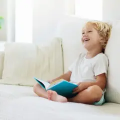 Chłopiec z uśmiechem na twarzy siedzi na kanapie z książką. To świetny sposób, jak nauczyć dziecko czytać, rozbudzając jego ciekawość.