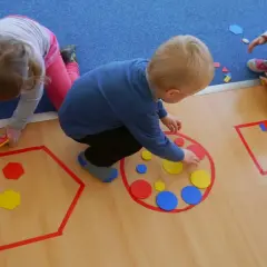 Dzieci w przedszkolu bawią się kolorowymi figurami geometrycznymi, tworząc wzory w wyznaczonych obszarach.
