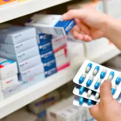 Ręce trzymają opakowania z kapsułkami, prawdopodobnie 3 dniowy antybiotyk na zapalenie ucha, na tle aptecznych półek z lekami.