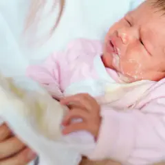 Noworodek w różowym pajacyku z widocznym ulewaniem. Mama wyciera buźkę dziecka.