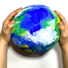 Dziecięca praca plastyczna przedstawiająca planetę Ziemię, namalowana farbami. Dłonie trzymają ją delikatnie, jakby chroniły nasz dom.