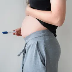 Kobieta w ciąży, trzymająca test ciążowy. Brzuszek widoczny, sugeruje 1 trymestr ciąży.