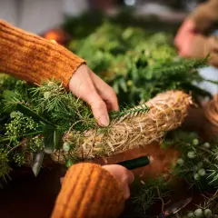 Ręce w pomarańczowym swetrze tworzą stroik z gałązek iglaków i słomy. Widać sznurek i świecę.