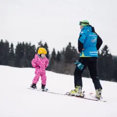 Mała dziewczynka w różowym kombinezonie i żółtym kasku uczy się jeździć na nartach pod okiem instruktora.