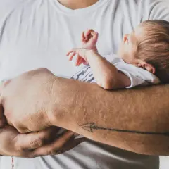 Ojciec tuli śpiącego niemowlaka. Maluch ma zamknięte oczy, a jego rączki delikatnie unoszą się w powietrzu. Tata z tatuażem strzały na przedramieniu czule obejmuje swoje dziecko.