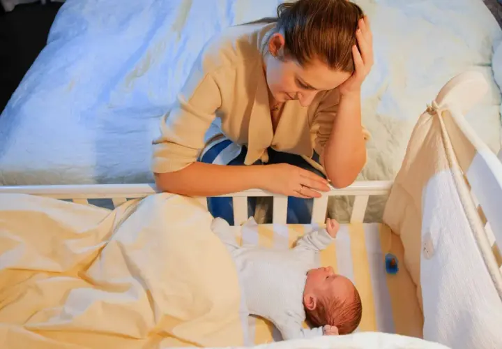 Zmęczona mama pochyla się nad łóżeczkiem, przeżywając kryzys snu u 9-miesięcznego dziecka.