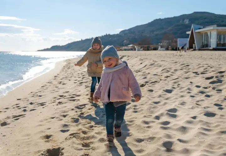 Dwa uśmiechnięte dzieci biegają po piaszczystej plaży. W tle widać morze i góry.