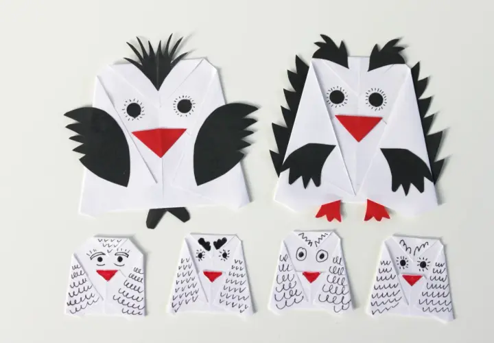 Dwie duże i cztery małe sowy z papieru, wykonane techniką origami. Każda sowa z papieru ma czerwony dziób i czarne detale.