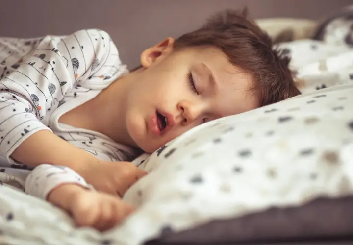 Małe dziecko śpi z otwartą buzią, wtulone w poduszkę. Jego sen jest głęboki i spokojny.