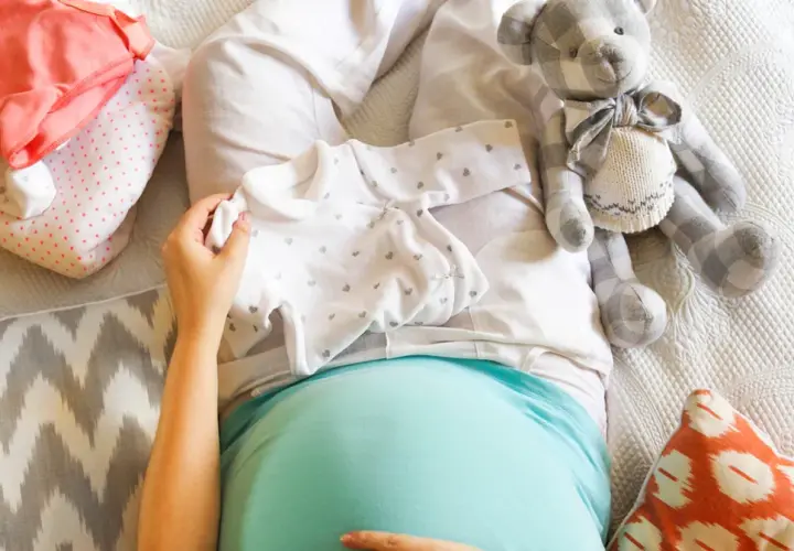Brzuszek w 34 tygodniu ciąży, przygotowania do narodzin: ubranka, pluszowy miś i poduszki.