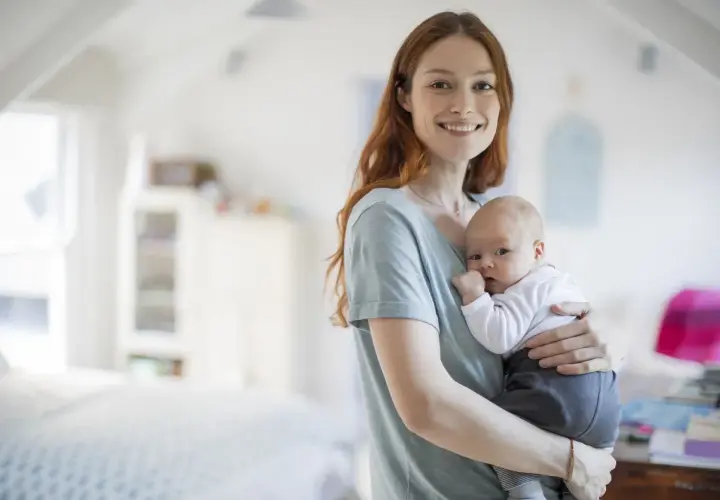 Uśmiechnięta mama trzyma na rękach noworodka. Dziecko ma lekko otwarte usta, jakby właśnie dostało czkawki.