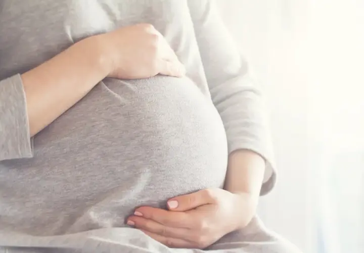 Brzuszek w 6 miesiącu ciąży, otulony szarym materiałem. Dłonie delikatnie go obejmują, czuć radość oczekiwania.