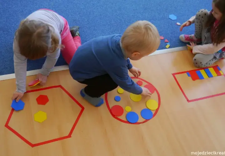 Dzieci w przedszkolu bawią się kolorowymi figurami geometrycznymi, tworząc wzory w wyznaczonych obszarach.