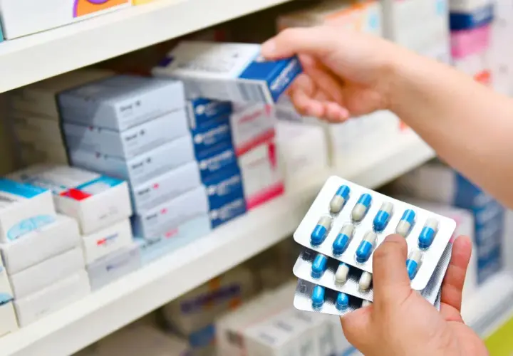 Ręce trzymają opakowania z kapsułkami, prawdopodobnie 3 dniowy antybiotyk na zapalenie ucha, na tle aptecznych półek z lekami.