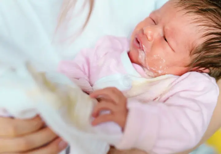 Noworodek w różowym pajacyku z widocznym ulewaniem. Mama wyciera buźkę dziecka.