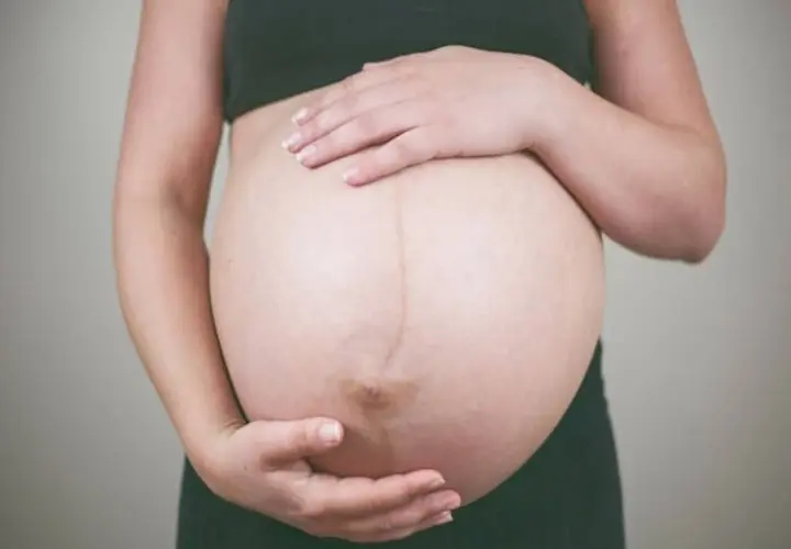 Dłońmi obejmuje zaokrąglony brzuch w ciąży. Widoczna ciemna linia biegnąca przez środek.