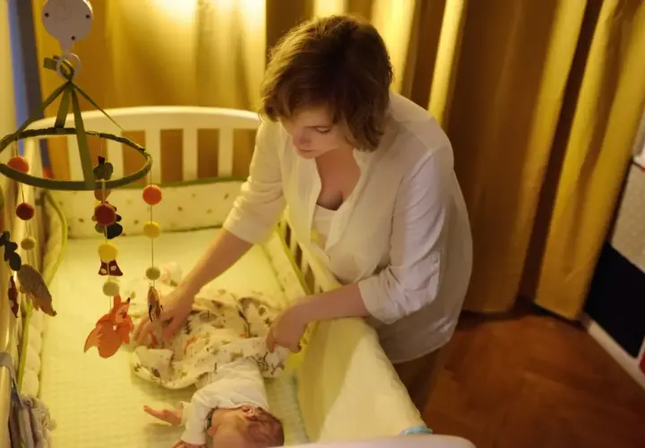 Mama uspokaja dziecko w łóżeczku, by złagodzić lęk separacyjny u niemowląt. Nad łóżeczkiem wisi karuzela z leśnymi zwierzątkami.