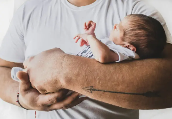 Ojciec tuli śpiącego niemowlaka. Maluch ma zamknięte oczy, a jego rączki delikatnie unoszą się w powietrzu. Tata z tatuażem strzały na przedramieniu czule obejmuje swoje dziecko.
