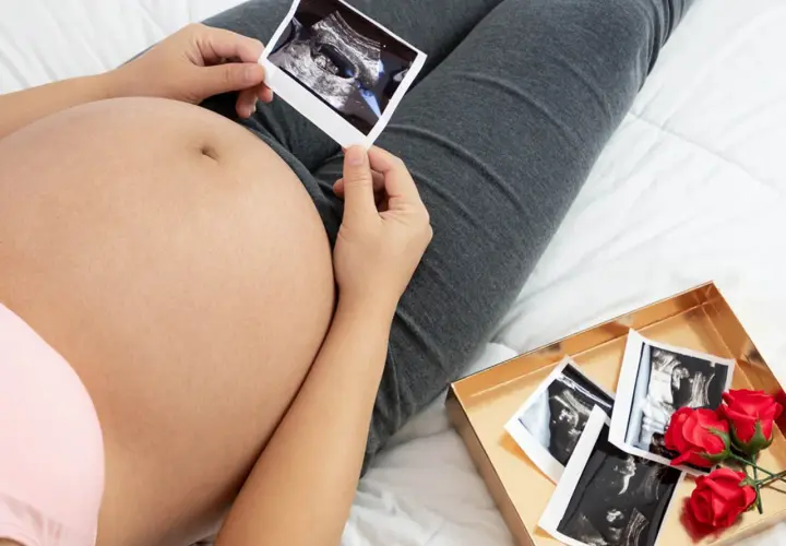 Kobieta trzyma zdjęcie USG na tle swojego brzucha ciążowego. Obok leży pudełko z kolejnymi zdjęciami i czerwonymi różami.