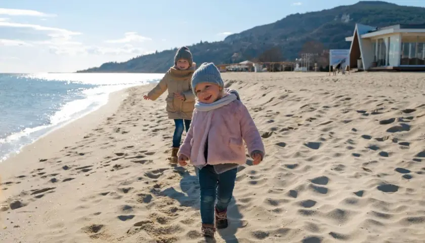 Dwa uśmiechnięte dzieci biegają po piaszczystej plaży. W tle widać morze i góry.