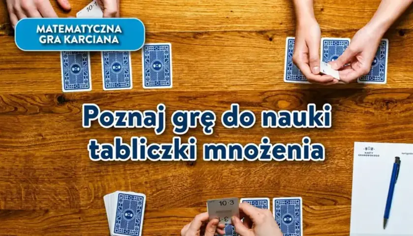 Gra karciana "Poznaj grę do nauki tabliczki mnożenia" na drewnianym stole. Dłonie trzymają karty z działaniami.