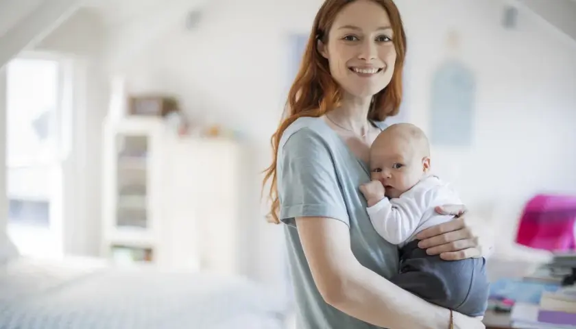Uśmiechnięta mama trzyma na rękach noworodka. Dziecko ma lekko otwarte usta, jakby właśnie dostało czkawki.