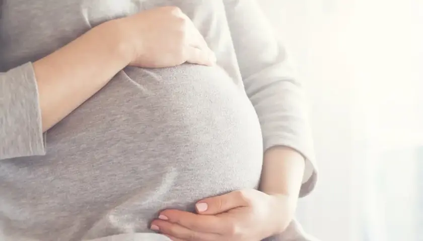 Brzuszek w 6 miesiącu ciąży, otulony szarym materiałem. Dłonie delikatnie go obejmują, czuć radość oczekiwania.
