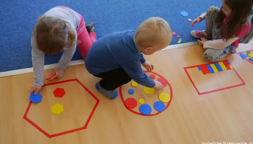 Dzieci w przedszkolu bawią się kolorowymi figurami geometrycznymi, tworząc wzory w wyznaczonych obszarach.