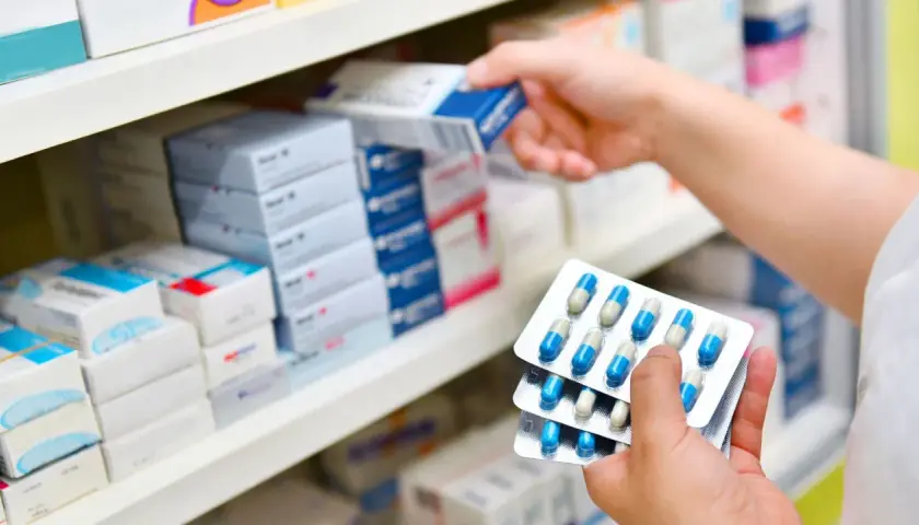 Ręce trzymają opakowania z kapsułkami, prawdopodobnie 3 dniowy antybiotyk na zapalenie ucha, na tle aptecznych półek z lekami.