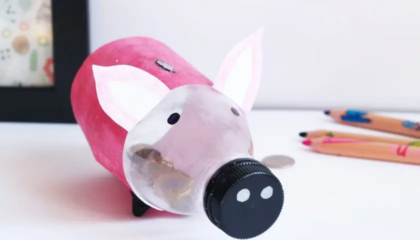 Urocza skarbonka diy z butelki, ozdobiona różowym papierem i uszami. W jej brzuszku widać monety.