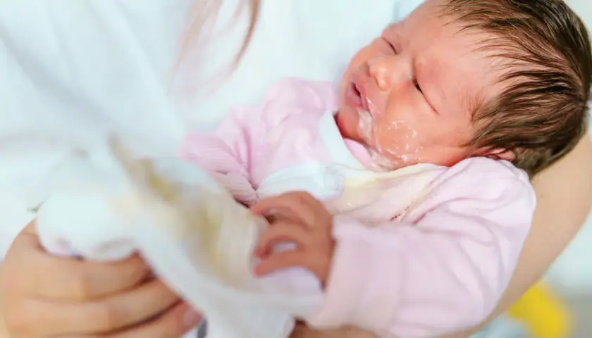 Noworodek w różowym pajacyku z widocznym ulewaniem. Mama wyciera buźkę dziecka.