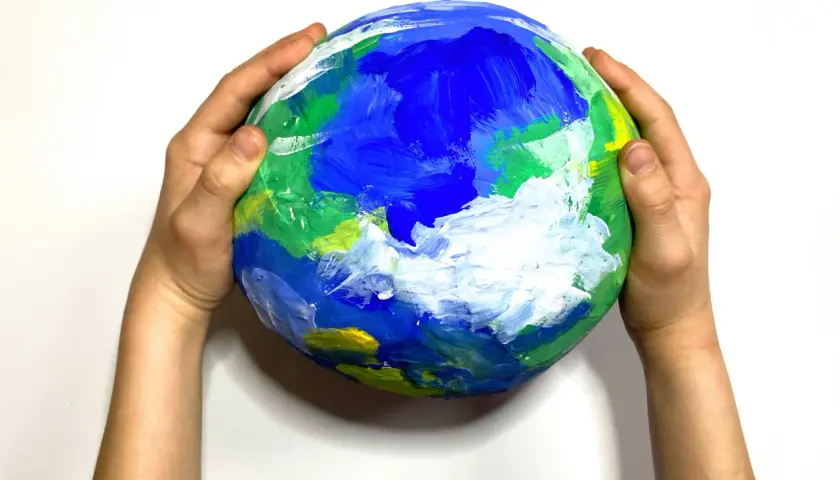 Dziecięca praca plastyczna przedstawiająca planetę Ziemię, namalowana farbami. Dłonie trzymają ją delikatnie, jakby chroniły nasz dom.
