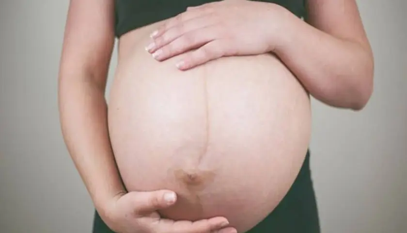 Dłońmi obejmuje zaokrąglony brzuch w ciąży. Widoczna ciemna linia biegnąca przez środek.
