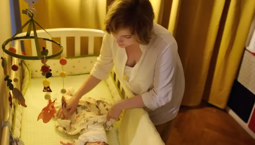 Mama uspokaja dziecko w łóżeczku, by złagodzić lęk separacyjny u niemowląt. Nad łóżeczkiem wisi karuzela z leśnymi zwierzątkami.
