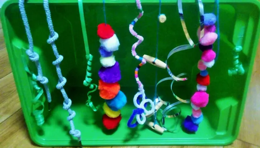 Kolorowe, własnoręcznie zrobione zabawki sensoryczne dla niemowląt diy: sznurki z supełkami, pompony, wstążki i rurki.