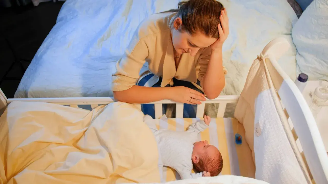 Zmęczona mama pochyla się nad łóżeczkiem, przeżywając kryzys snu u 9-miesięcznego dziecka.