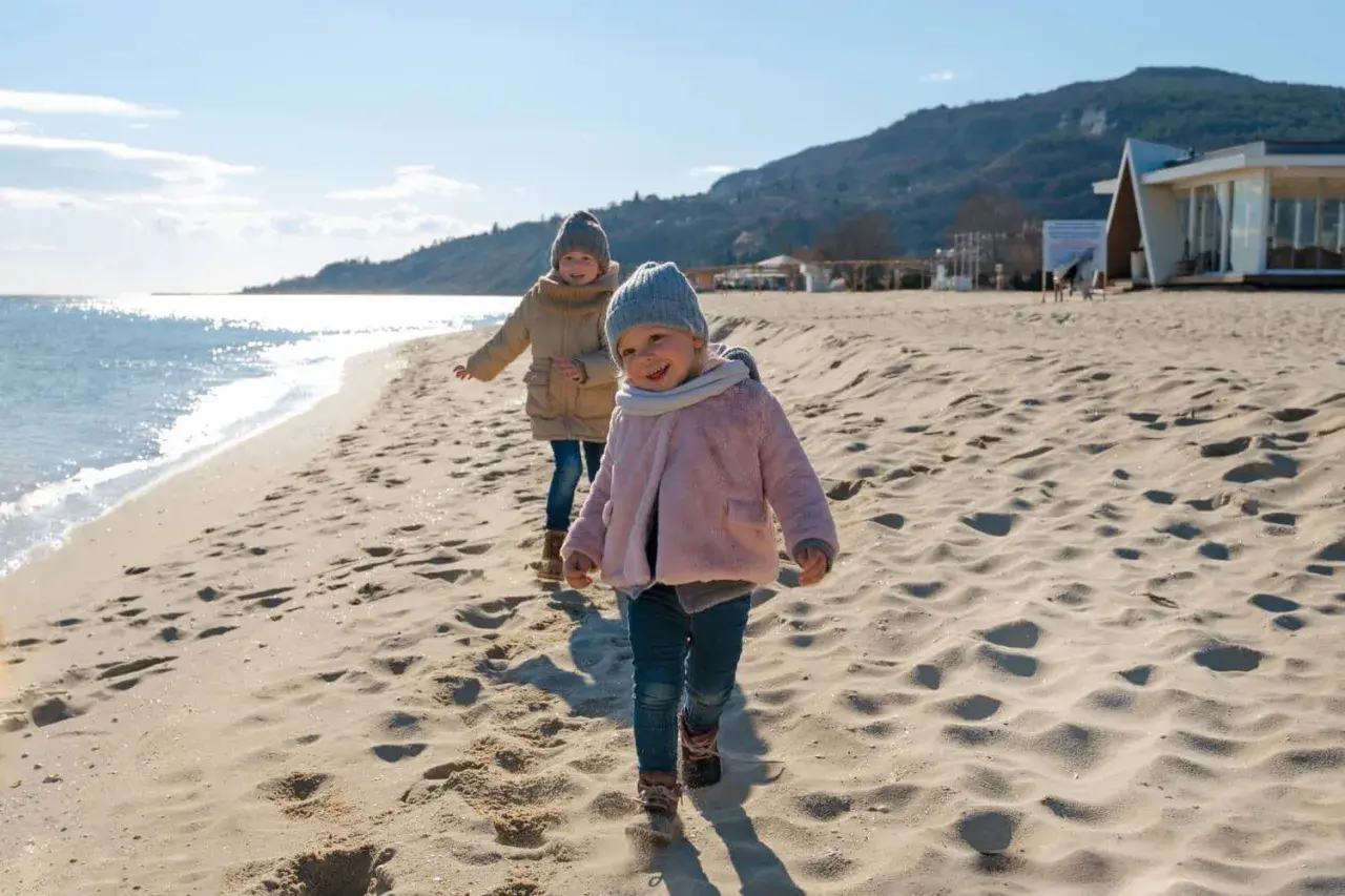 Dwa uśmiechnięte dzieci biegają po piaszczystej plaży. W tle widać morze i góry.
