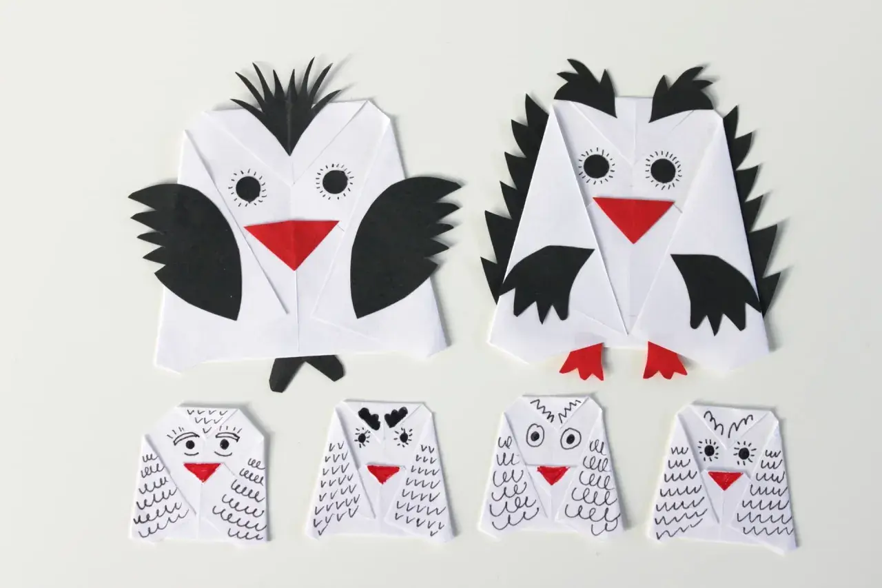 Dwie duże i cztery małe sowy z papieru, wykonane techniką origami. Każda sowa z papieru ma czerwony dziób i czarne detale.
