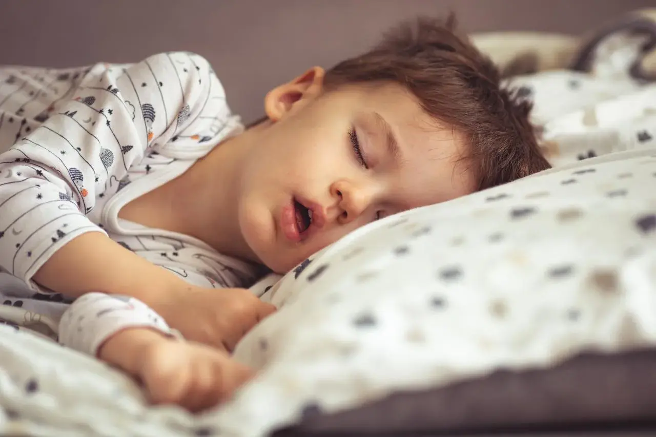 Małe dziecko śpi z otwartą buzią, wtulone w poduszkę. Jego sen jest głęboki i spokojny.