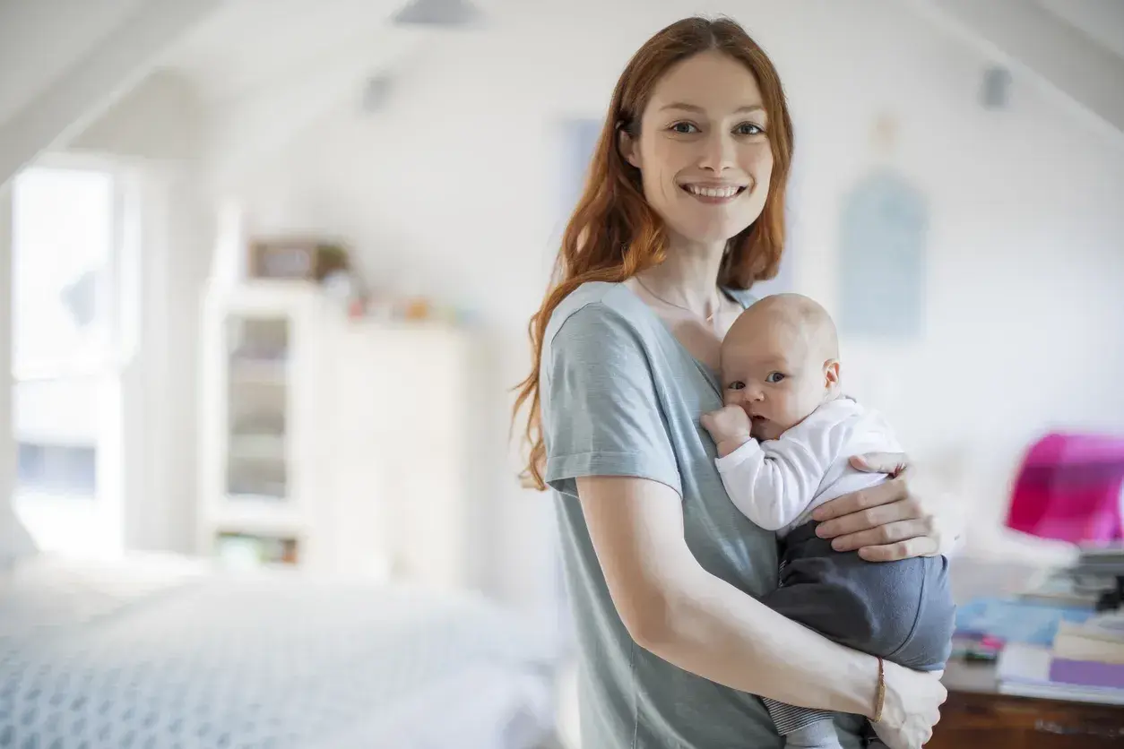 Uśmiechnięta mama trzyma na rękach noworodka. Dziecko ma lekko otwarte usta, jakby właśnie dostało czkawki.