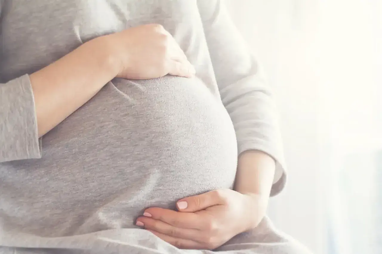 Brzuszek w 6 miesiącu ciąży, otulony szarym materiałem. Dłonie delikatnie go obejmują, czuć radość oczekiwania.