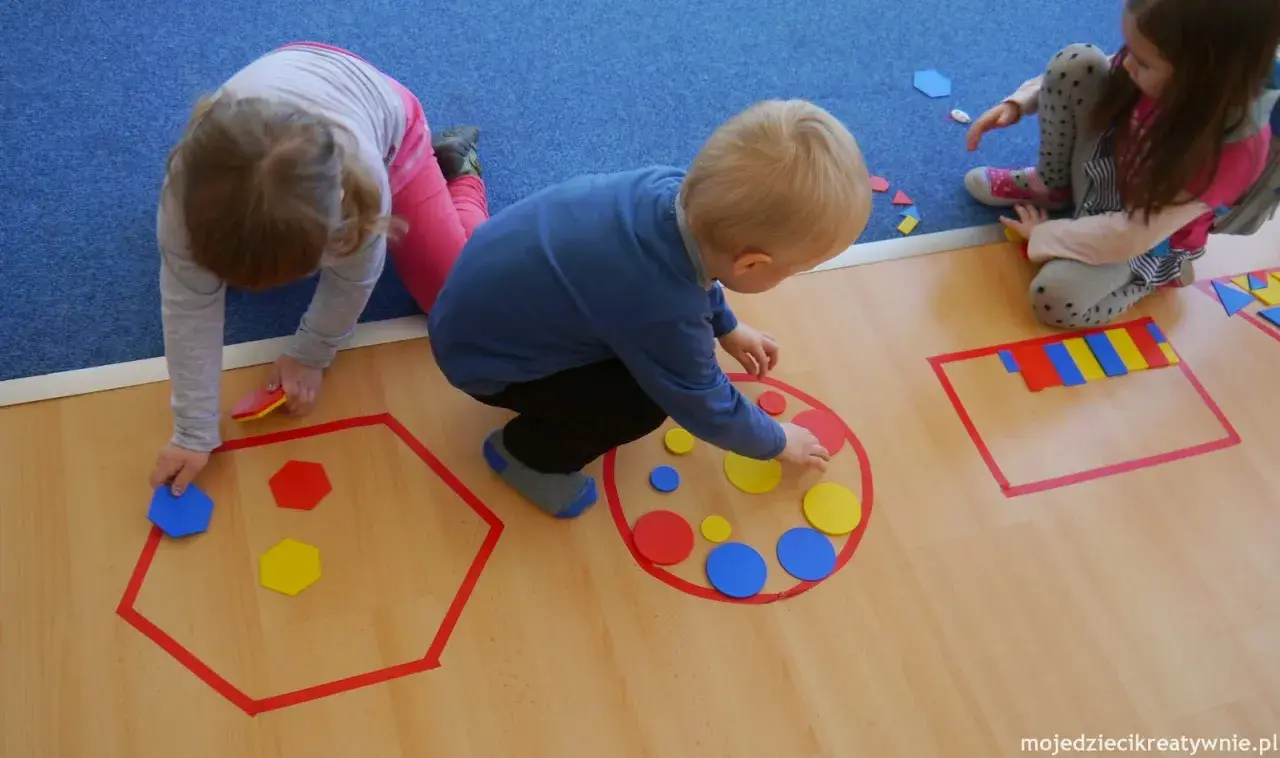 Dzieci w przedszkolu bawią się kolorowymi figurami geometrycznymi, tworząc wzory w wyznaczonych obszarach.